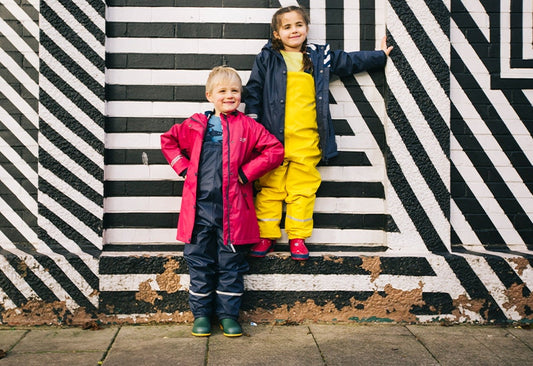 children stood against black and white striped wall, wearing vibrant muddy puddles waterproofs