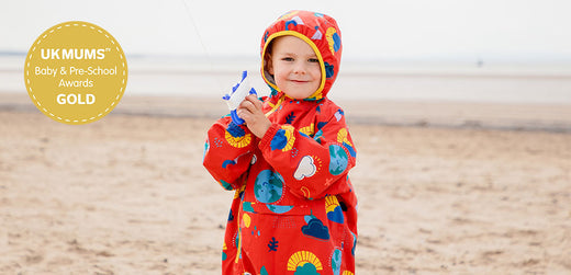 Child wearing colorful waterproof raincoat with hood on a sandy beach, holding a kite. UK Mums awards gold badge visible.