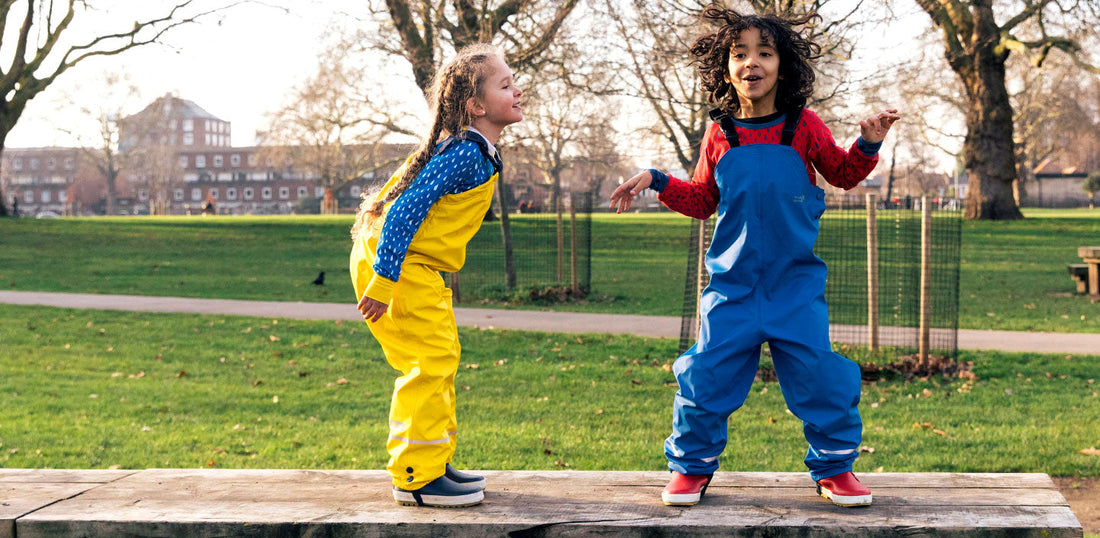 Children playing outdoors in waterproof overalls, one in yellow, one in blue, in a park