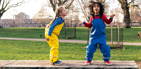 Children playing outdoors in waterproof overalls, one in yellow, one in blue, in a park