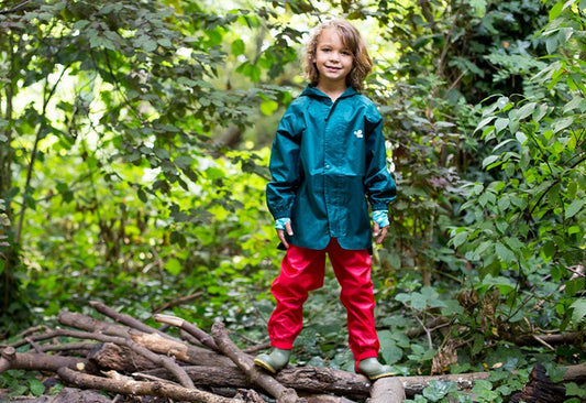 boy stood on tree branch, surrounded by trees, wearing muddy puddles waterproofs