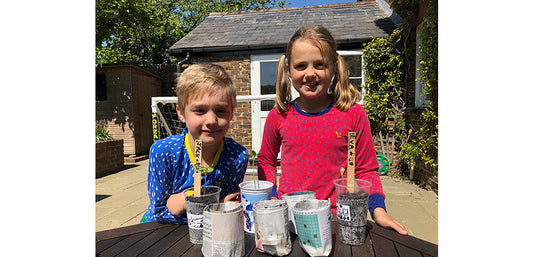 boy and girl stood in front of garden table with handmade newspaper plant pots, wearing vibrant muddy puddles thermals
