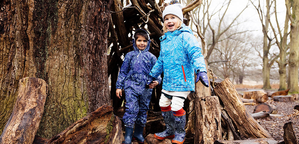 Two children in a wooded area wearing durable, waterproof gear, including a patterned coat, hat, and snowsuit, near a teepee structure of branches.