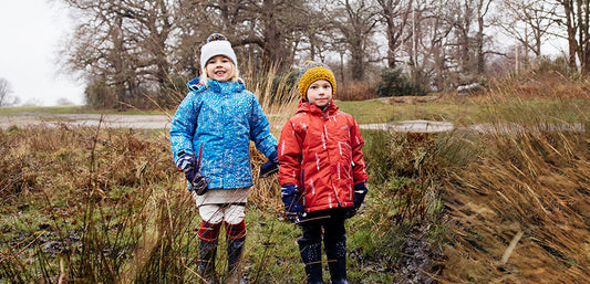 boy and girl stood on grass surrounded by trees, wearing muddy puddles ski jackets and bobble hats