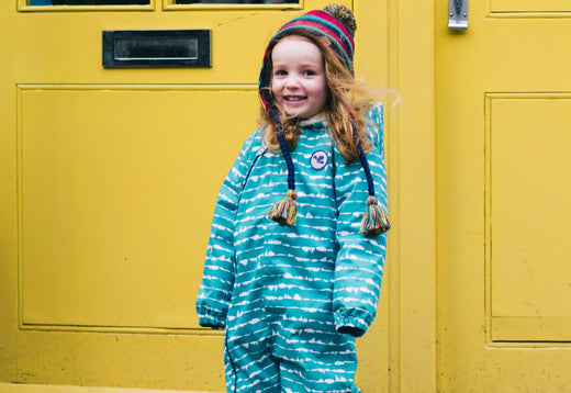 girl stood in front of yellow door, wearing green muddy puddles puddlesuit and trapper hat