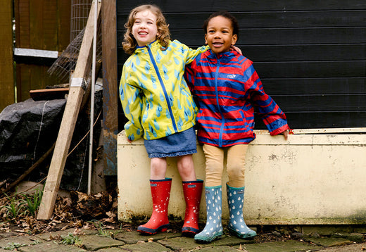 two girls stood in front of metal trough, wearing colourful patterned muddy puddles waterproof jackets and wellies