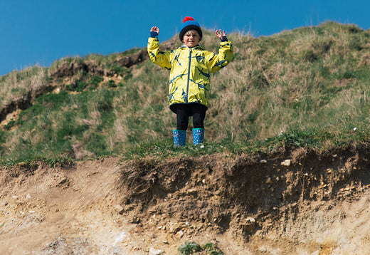 boy stood on grassy mound wearing vibrant yellow waterproof jacket, blue wellies and bobble hat