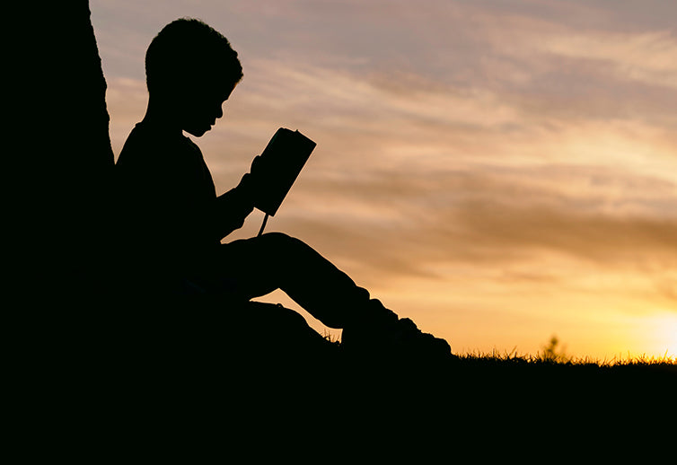 silhouette of child sat at base of tree reading a book, with sunset in the background