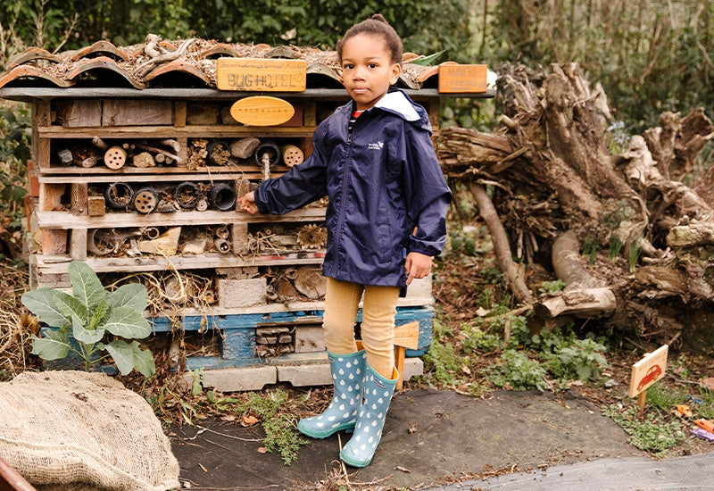 girl stood in front of large bug hotel, wearing muddy puddles waterproof jacket, thermals and wellies