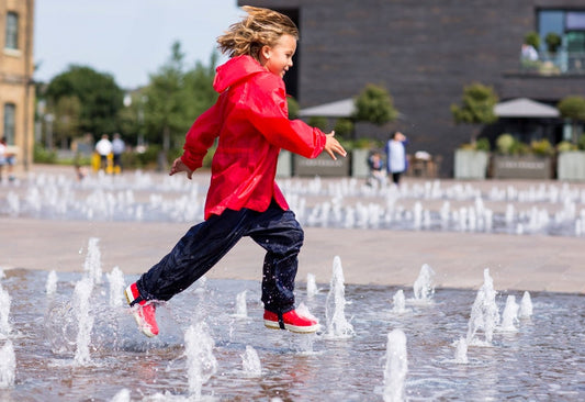 child running through water fountains wearing muddy puddles waterproof jacket, trousers and wellies 