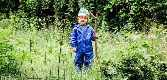 boy standing in grassy area holding large stick, with feather crown and wearing muddy puddles originals waterproof puddlesuit