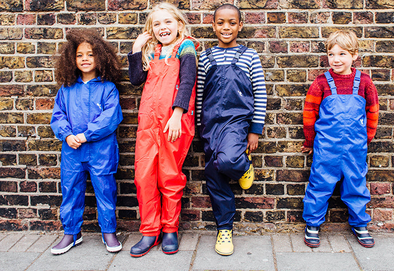 children stood in front of brick wall, wearing vibrant muddy puddles waterproof dungarees and puddlesuit