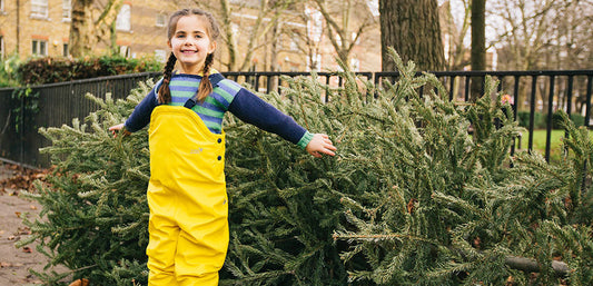 girl stood in front of christmas trees wearing yellow waterproof dungarees and blue striped jumper