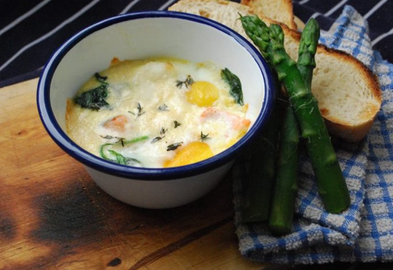 eggs en cocotte in blue and white enamel bowl, next to blue and white tea towel, crusty bread and asparagus