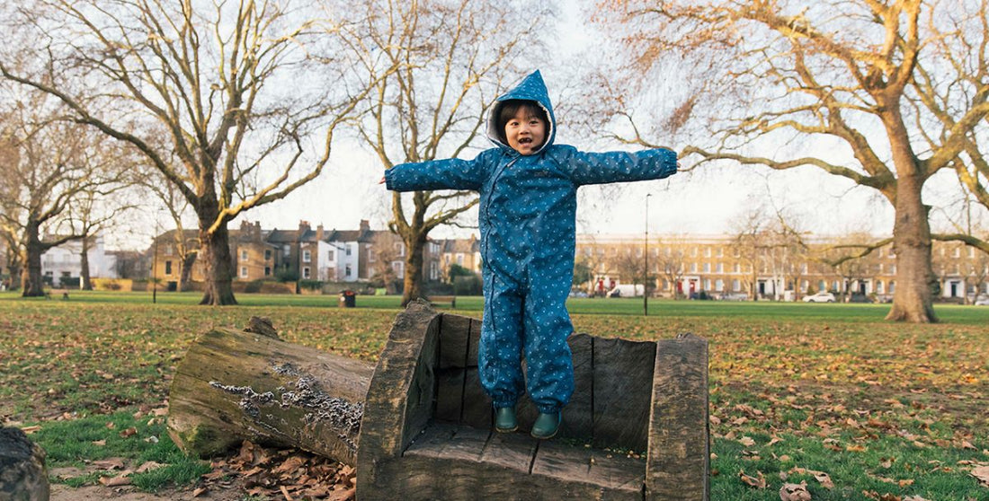 child jumping on a woodland park bench, wearing a turquoise muddy puddles scampsuit, with trees and houses in the background