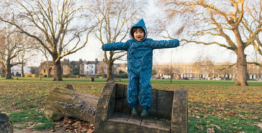 child jumping on a woodland park bench, wearing a turquoise muddy puddles scampsuit, with trees and houses in the background