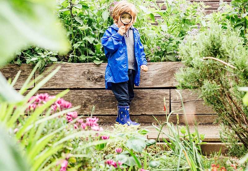 boy stood in garden holding magnifying glass and wearing vibrant blue waterproof jacket and wellies