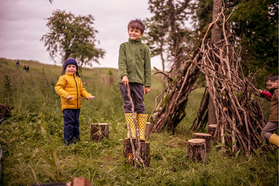 children stood in a grassy woodland area near a handmade twig den