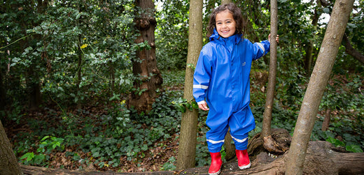 Child in blue waterproof rain suit and red boots standing on a log in a forest