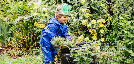 Child in blue waterproof suit gardening outside surrounded by lush plants and yellow flowers.
