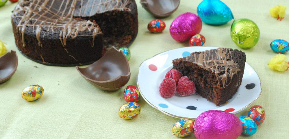 Chocolate cake with a slice on a white dish, drizzled with chocolate, accompanied by raspberries, chocolate shells, and Easter-themed foil-wrapped chocolate eggs.