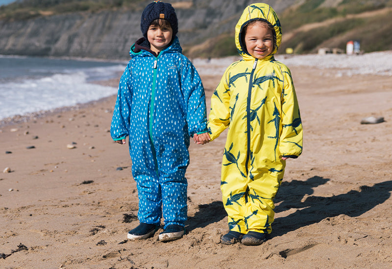 Two children in durable, waterproof suits—one blue with raindrops, one yellow with sharks—stand smiling on a beach, embodying Muddy Puddles' outdoor spirit.