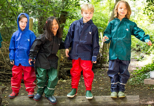 children stood on log in forest school setting, wearing colourful muddy puddles waterproof jackets and trousers