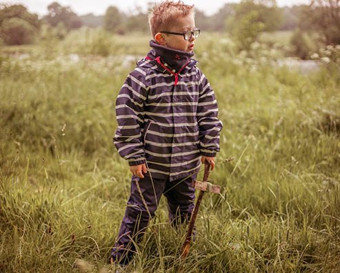 boy standing in long grass holding a long stick, wearing a striped muddy puddles waterproof jacket