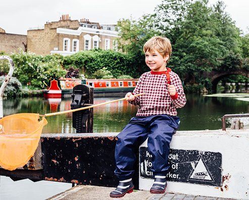 boy sitting by a canal holding a fishing net, wearing muddy puddles waterproof trousers and wellies