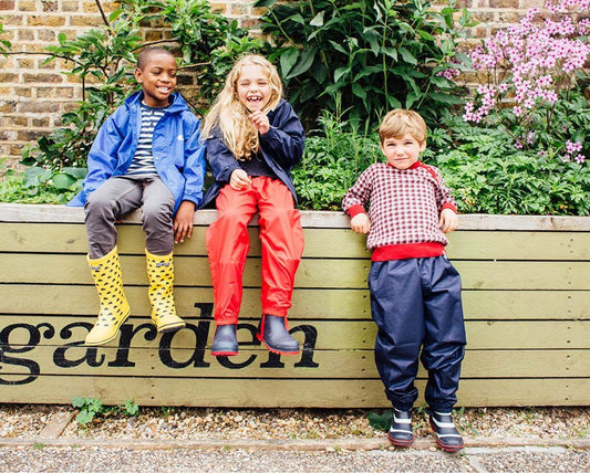 children sitting on and standing in front of a garden wall, wearing vibrant muddy puddles raincoats, waterproof trousers and wellies
