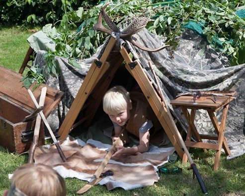 boys playing with an outdoor wooden den covered in tarpaulin and leaves