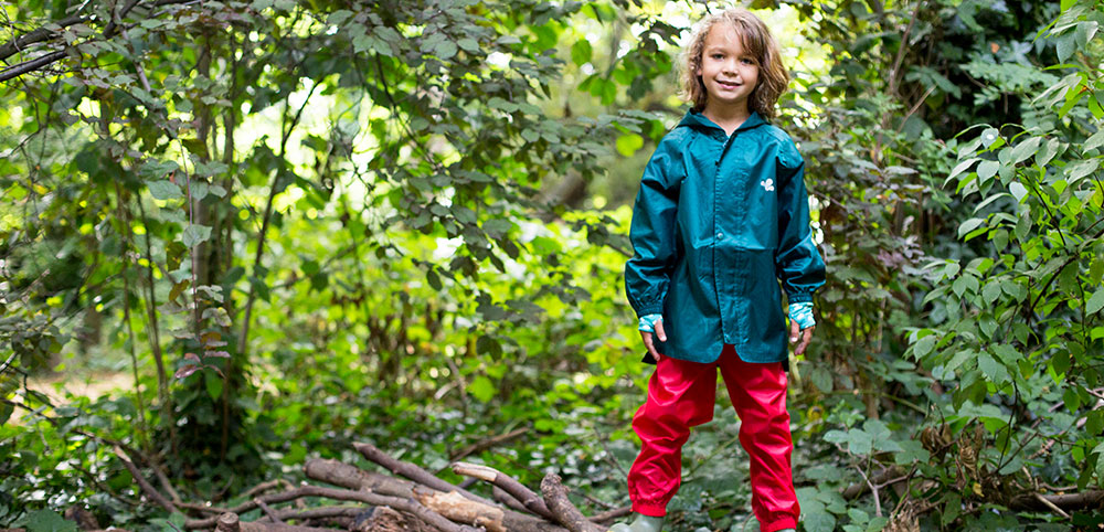 boy stood in woodland setting wearing muddy puddles green waterproof jacket and red waterproof trousers