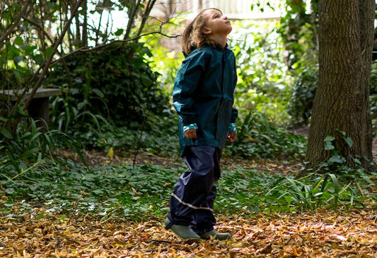 child stood in forest clearing looking up at the trees, wearing muddy puddles green waterproof jacket and navy trousers