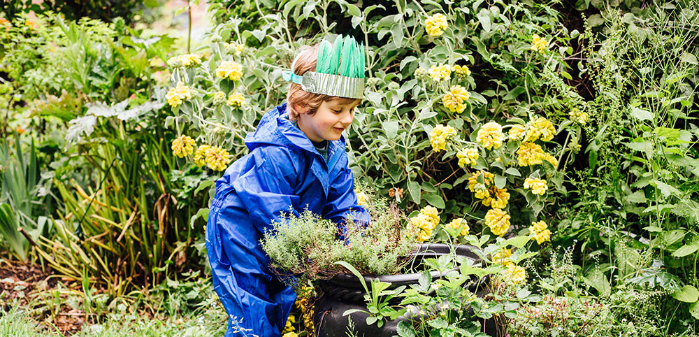 young boy gardening wearing muddy puddles blue puddle suit and feather crown