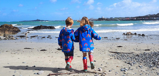 Two children in blue rainbow waterproof jackets and red boots walking on a windy beach