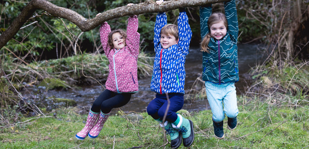 children hanging from tree branch wearing colourful muddy puddles waterproof jackets and wellies
