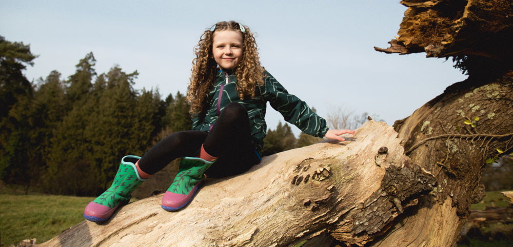 Child in patterned waterproof jacket and green wellington boots sitting on a fallen tree outdoors