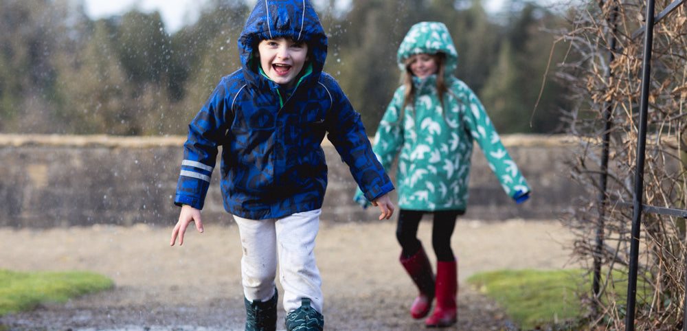 children running through rain wearing muddy puddles waterproof ski jackets, with brick wall and trees behind them