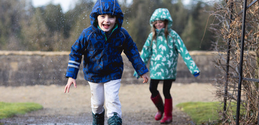 children running through rain wearing muddy puddles waterproof ski jackets, with brick wall and trees behind them