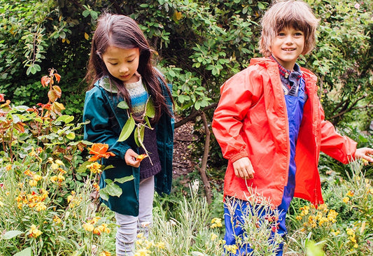 girl and boy standing in woodland garden looking at plants, wearing vibrant muddy puddles waterproof jackets