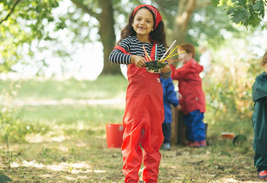 girl at forest school setting holding mud pie, wearing red muddy puddles waterproof dungarees and matching bandana