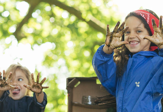 children at forest school holding up muddy hands to camera, wearing blue muddy puddles puddlesuits