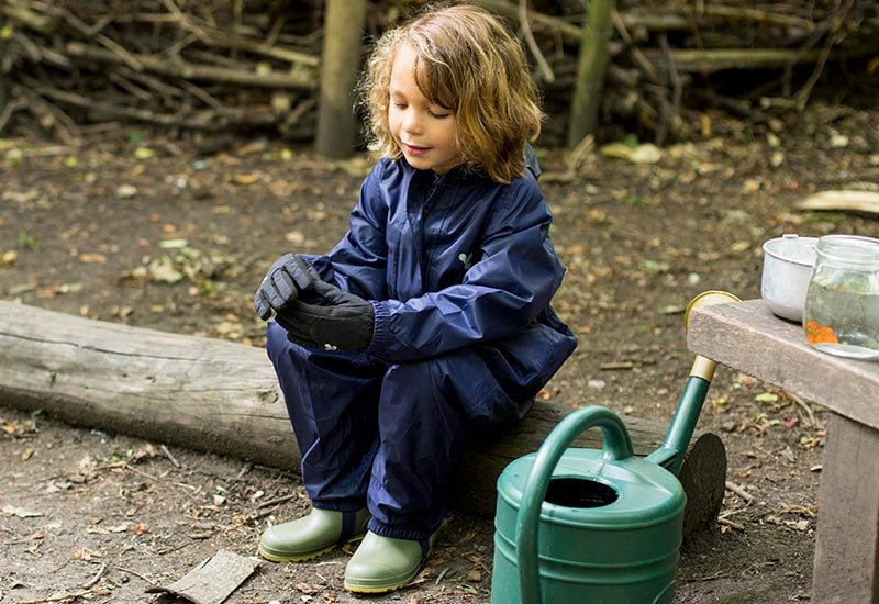 child sitting on log next to watering can, wearing navy muddy puddles puddlesuit