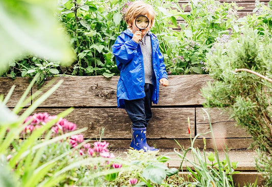 boy stood in garden holding magnifying glass and wearing blue muddy puddles waterproof jacket and wellies