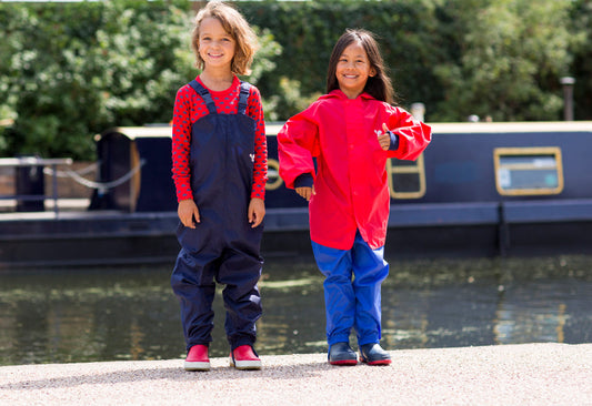 boy and girl standing by canal, wearing vibrant muddy puddles waterproof dungarees, jacket and trousers