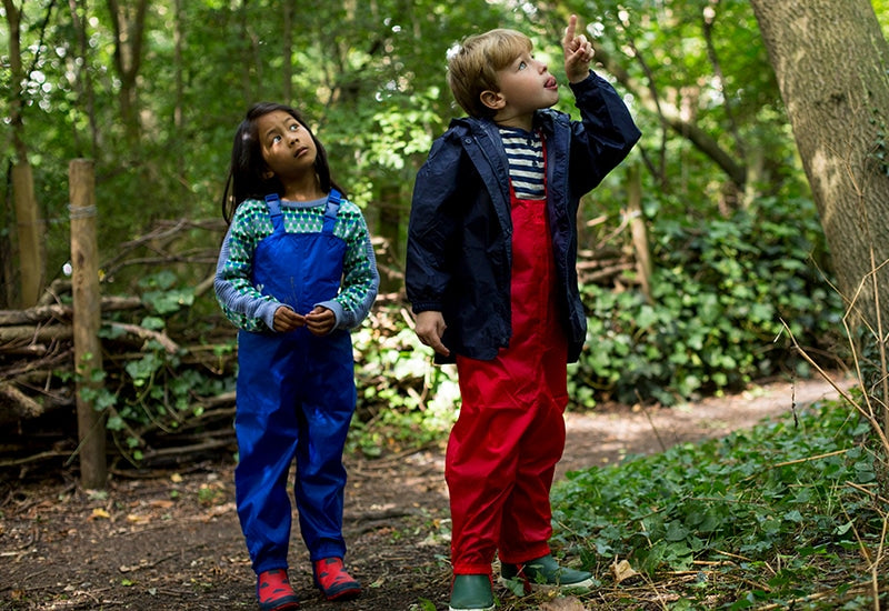 boy and girl stood in forest, looking up to treetops, wearing vibrant red and blue muddy puddles waterproofs