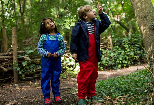 boy and girl stood in forest, looking up to treetops, wearing vibrant red and blue muddy puddles waterproofs