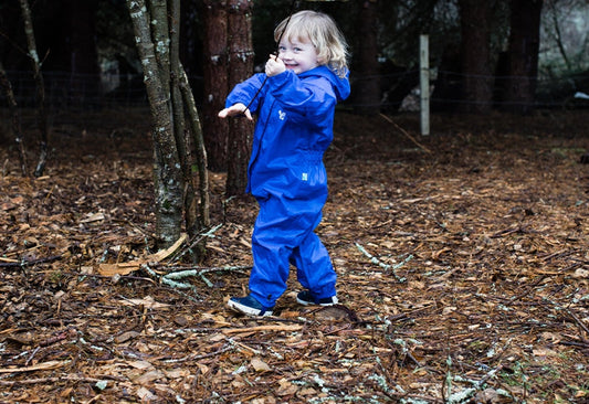 toddler standing in woodland holding a stick, wearing blue muddy puddles puddlesuit 