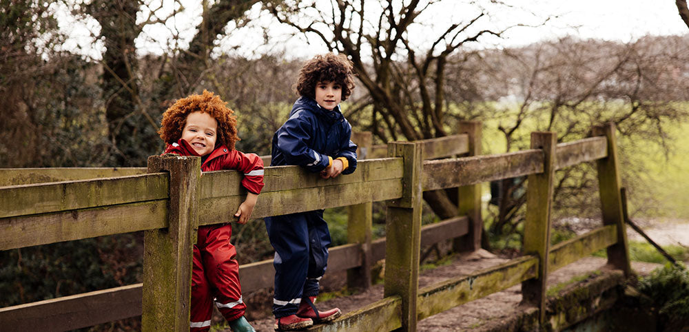 two boys stood on bridge wearing muddy puddles red and navy puddle suits
