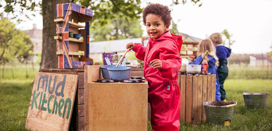 toddler boy playing in outdoor mud kitchen, wearing muddy puddles red puddlesuit